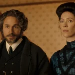 "A man with curly hair and a beard stands beside a woman with dark hair in an old-fashioned, elegant dress, both in a warmly lit vintage interior with patterned wallpaper and a staircase in the background."