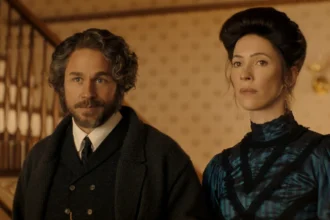 "A man with curly hair and a beard stands beside a woman with dark hair in an old-fashioned, elegant dress, both in a warmly lit vintage interior with patterned wallpaper and a staircase in the background."