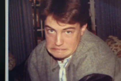 "Young man indoors making a humorous, exaggerated face, wearing a grey jacket with leather sleeves. The environment looks like a living room with curtains and patterned furniture in the background. Warm, casual setting."