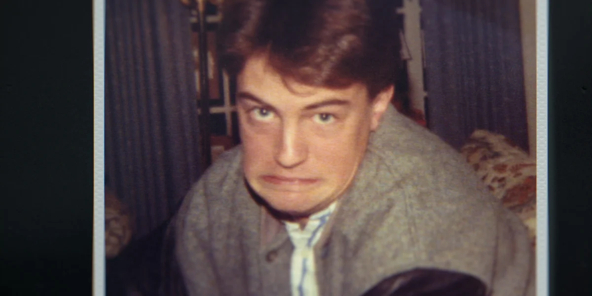 "Young man indoors making a humorous, exaggerated face, wearing a grey jacket with leather sleeves. The environment looks like a living room with curtains and patterned furniture in the background. Warm, casual setting."